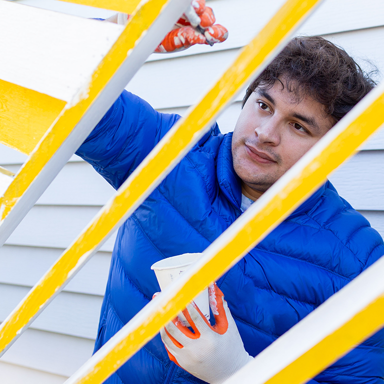 A college-aged boy wearing with dark curly hair and wearing a large puffy blue jacket, paints a bright yellow porch railing with white paint.  