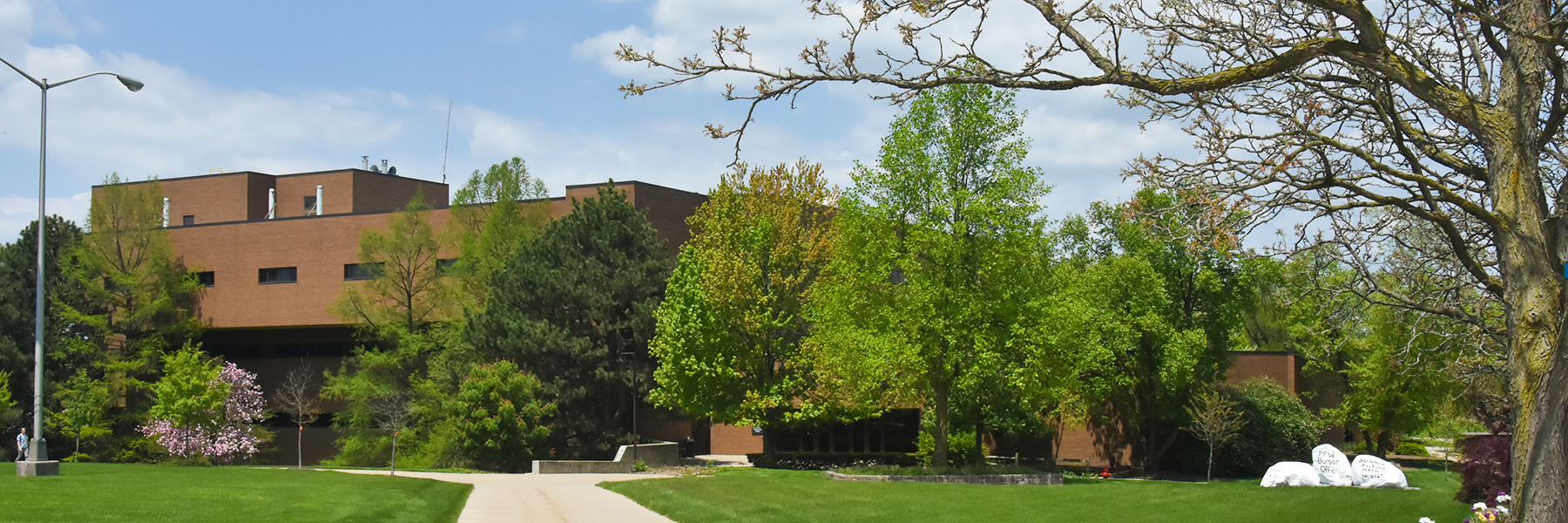 It's spring time at Indiana University Fort Wayne. The grass is green and some of the trees have flowers on them. Surrounding the lush landscape is Neff Hall. 